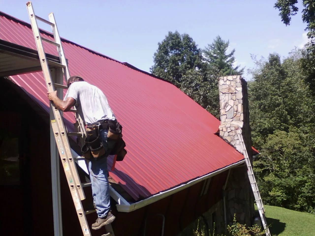 Roofer on a ladder beside a red metal roof and stone chimney — Soffit & Fascia Repair by Roofing Brothers in Ellsworth